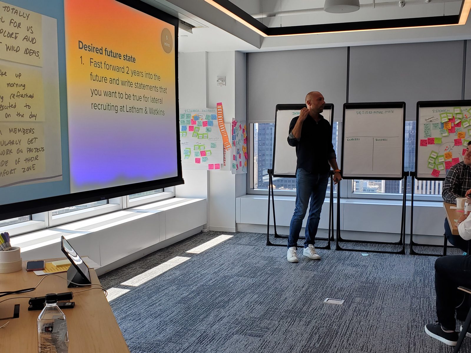 Joe Lalley leading a workshop, with a screen reading 'Desired future state' and sticky-note boards behind him